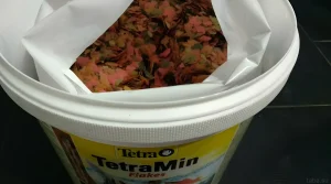 Фотография открытой банки корма TetraMin Flakes. Разноцветные хлопья, готовые к кормлению аквариумных рыбок, обеспечивают сбалансированное питание.